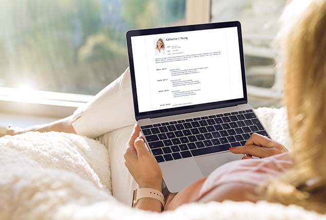 woman sits on white bed and looks at her resume on laptop