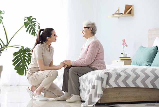 woman kneels talking to older woman sitting on foot of bed in white bedroom