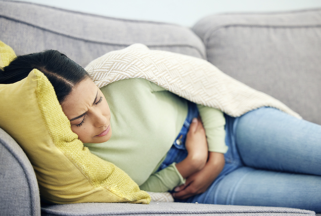 A woman lays on the sofa with a blanket over her as she holds her stomach in pain.