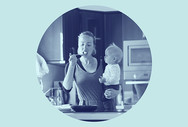 woman holds baby on her hip standing in a kitchen and eats food with a fork