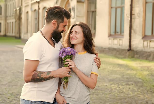 man and woman embrace in courtyard as they walk, he hands her a bouquet of purple flowers