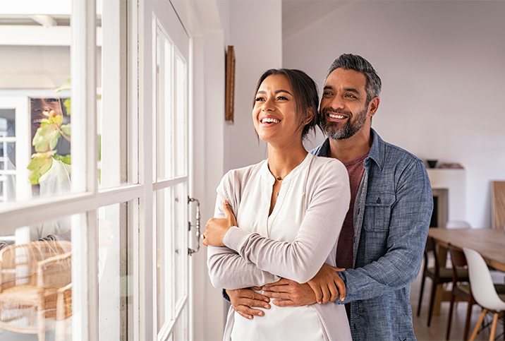 man and woman smile at sunny window as he holds her from behind