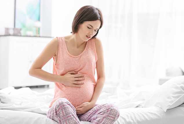 pregnant woman in pink tank top grabbing her stomach in pain while sitting on bed