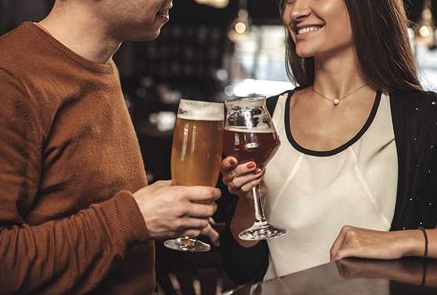 Two people smile at each other while holding glasses of beer at a bar.