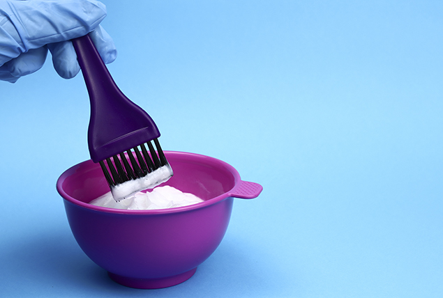 A gloved hand prepares hair bleach in a purple bowl.