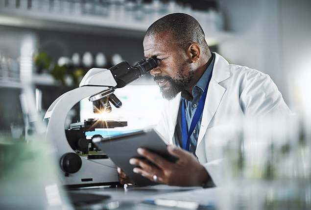 Black doctor in white lab coat looking into eyepiece of microscope holding tablet