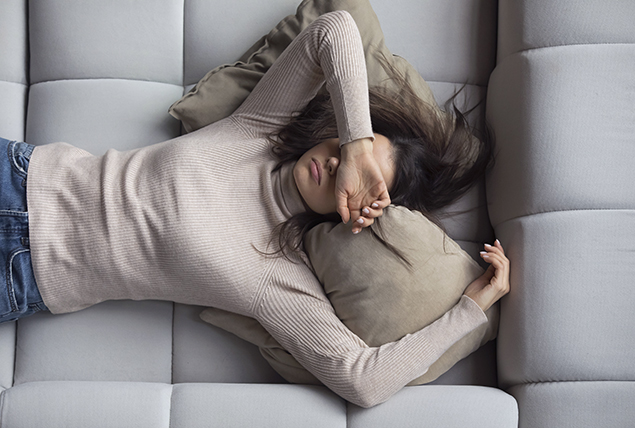 A tired woman lays on a couch with her arm over her face.
