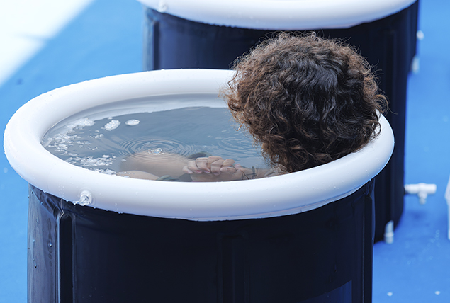 A woman sits in a deep tub doing an ice plunge with water up to her neck.
