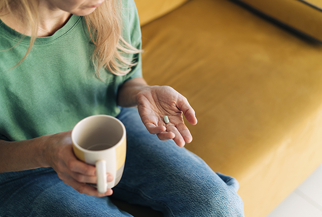 A woman holds a pill in her hand with the other hand holding a cup of coffee.