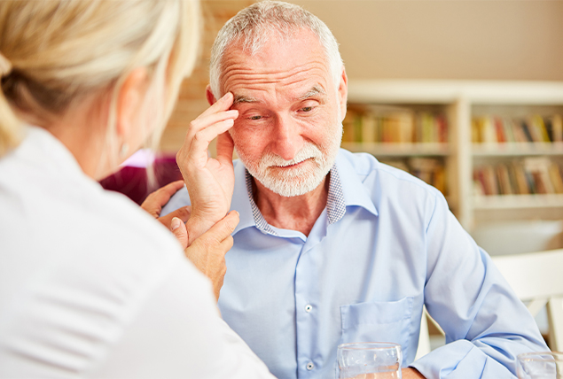 older man with white beard touching his temple while talking to blonde woman