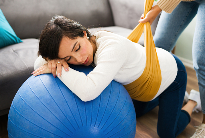 pregnant woman leaning on blue yoga ball with someone wrapping a yellow band around her belly
