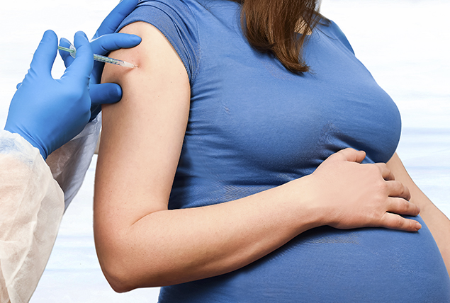 A pregnant woman in a blue shirt holds her stomach as she receives a vaccine in her right arm.