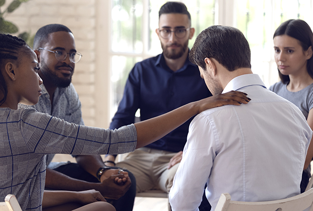 Four people sit with a man in a support group as a woman puts her hand on his shoulder.