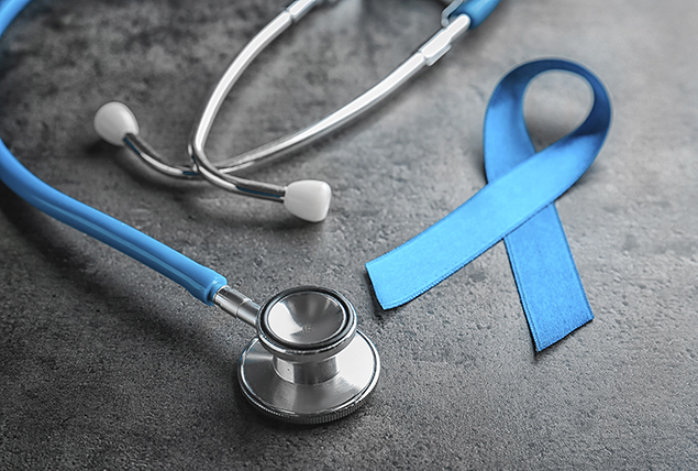 A blue prostate cancer ribbon sits beside a stethoscope against a grey surface.