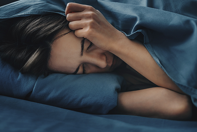 A woman lies in bed with her hand pressing her face into the pillow.