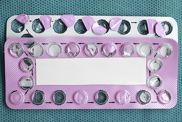 Two empty packets of birth control pills lie back to back against a dark teal background.