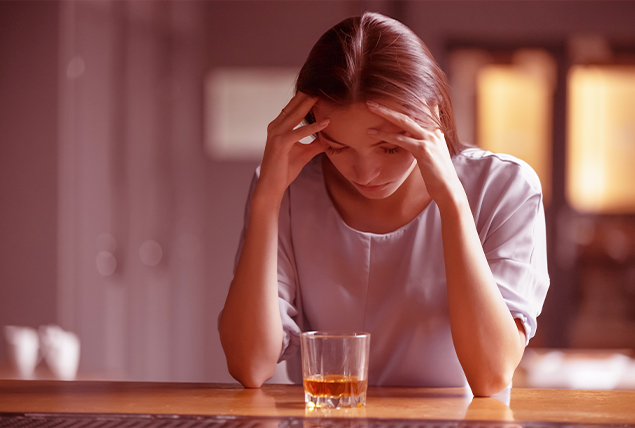 A woman holds her head between her hands as she looks down at a glass of whiskey.