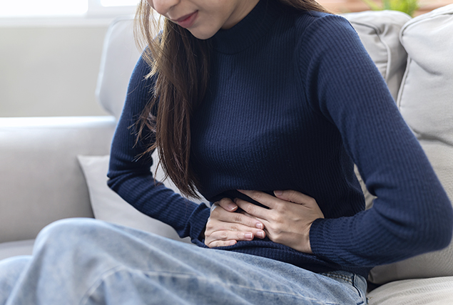 A woman holds her stomach in pain while sitting on the sofa.