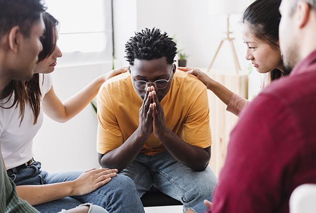 A man sits in the middle of a support group as people place their hands on his shoulders.