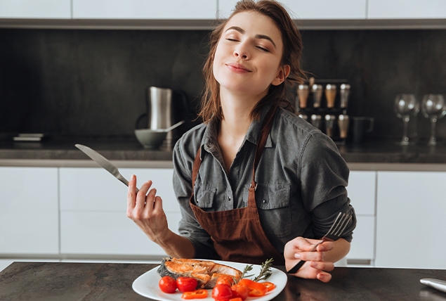 A woman sits in front of her plate with a knife and fork in hand and her eyes closed.