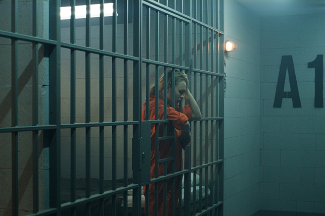 A woman in an orange jumpsuit sits inside of a jail cell hanging her arms over the bars.