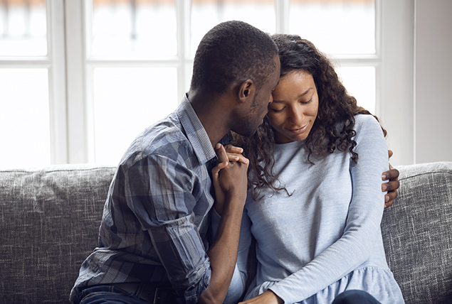 A man wraps his arm around a woman and holds her hand while sitting on a sofa.