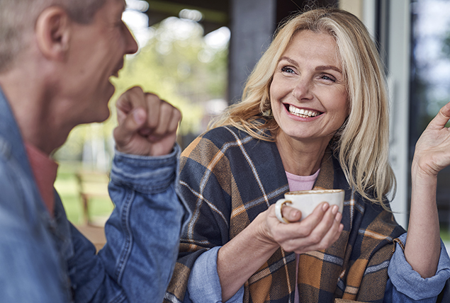 A woman holds a coffee cup in her hand as she smiles and talks to the man next to her.