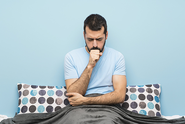 A man sits in bed under the covers coughing into his hand.