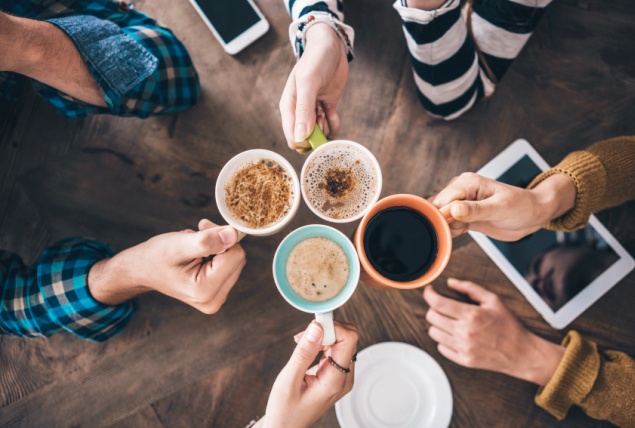 Four cups of coffee are held up together as people cheers over the table.