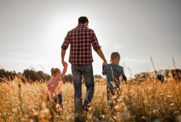 A father walks through a field holding hands with his two children.