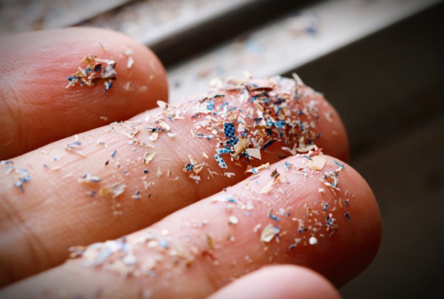 A close up photograph shows small pieces of microplastics held up on a set of fingers.