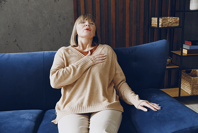 A woman sits on a sofa with her hand over her heart as she breathes slowly.