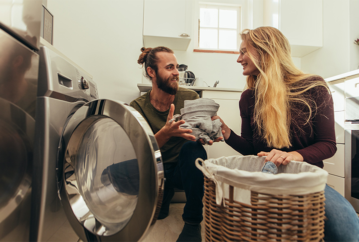 A man takes laundry out of the dryer and hands it to his female partner with a smile.