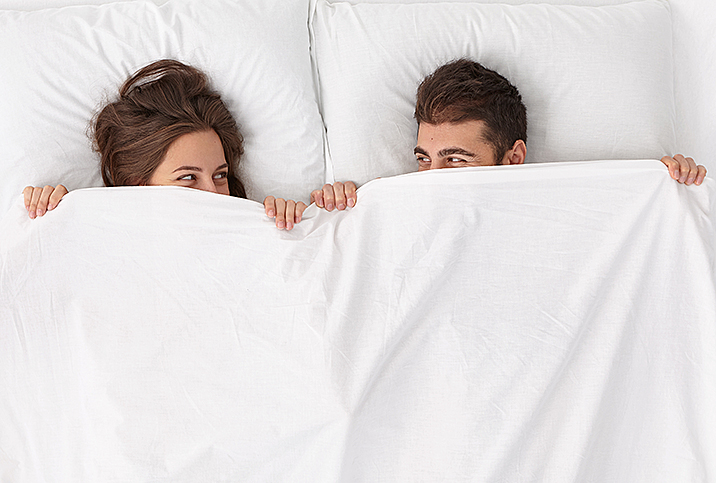 A man and woman lay beside each other in bed and hold the bedsheet up over their smiling faces.