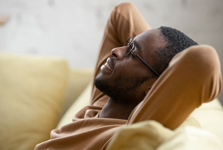 A black man leans back onto a sofa with his arms up behind his head.