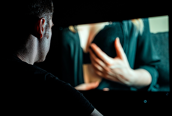 A man watches his computer screen where a woman is undressing and touching herself sexually.