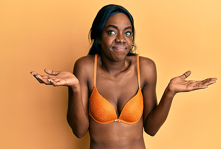 A black woman in an orange bra holds her arms up as she shrugs.