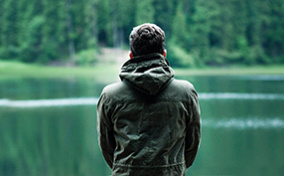 A man sits facing towards a lake outside in the forest.