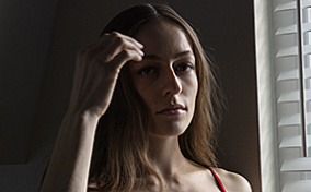 A woman in lingerie stands near a window with her hand brushing up near her hair.