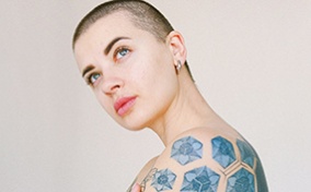 A tattooed person with a buzzcut crosses their arms and looks up thoughtfully.