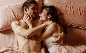 A couple lays in bed smiling at each other.