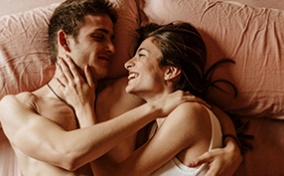 A couple lays in bed smiling at each other.