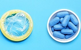 Blue pills sit in a small open white container next to a condom on a blue surface.