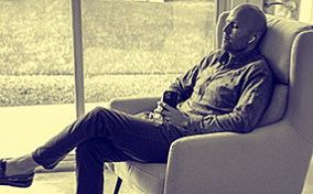 A man sits in a chair with earbuds in listening to something playing on his cellphone.