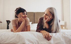 A mom and daughter lay on a bed smiling at each other.