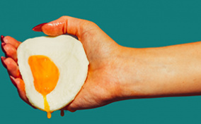 The hand of a woman holds a fried egg with yolk dripping down against a green background.