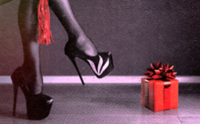 A dominatrix holds a red whip over her legs in platform high heels with a red gift nearby.