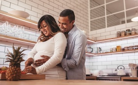 A man hugs a pregnant woman from behind while she cooks in the kitchen.