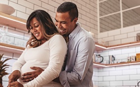 A man hugs a pregnant woman from behind while she cooks in the kitchen.