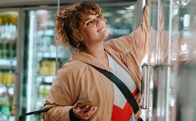 A woman is grocery shopping and reaching into the freezer.